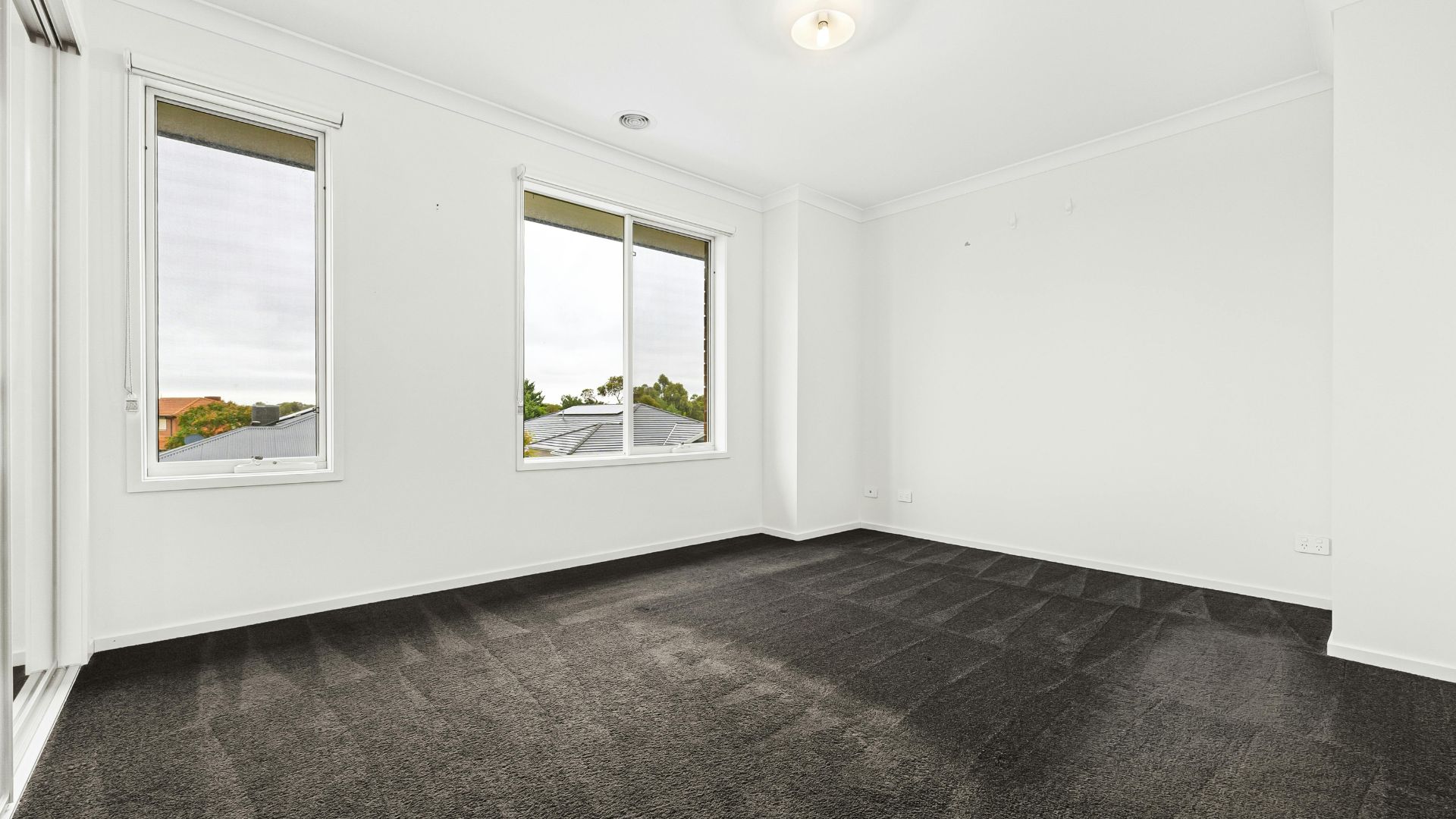 Empty room with white walls, dark carpet, and two windows with outside view